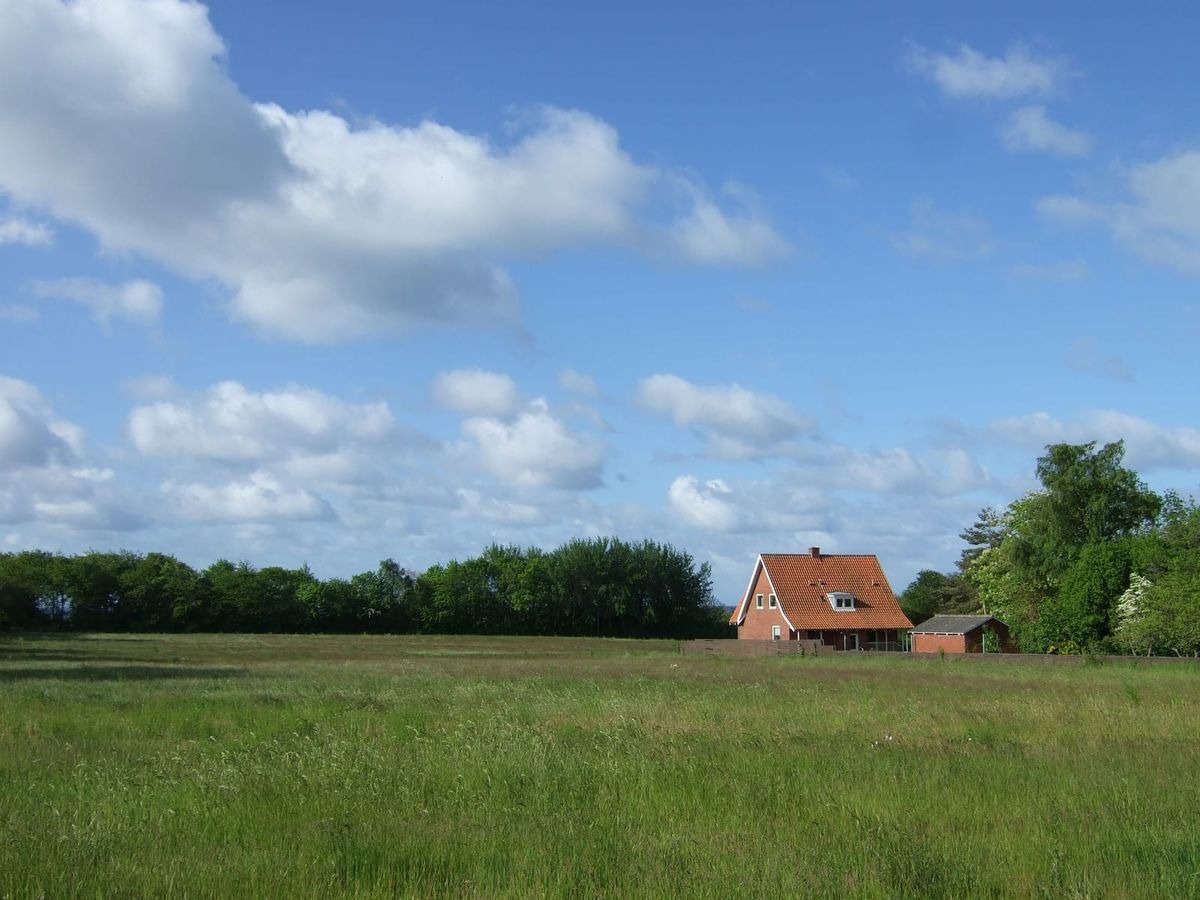 Maison en briques au toit de tuiles rouges, au fond d'un grand pré, sous un ciel nuageux.